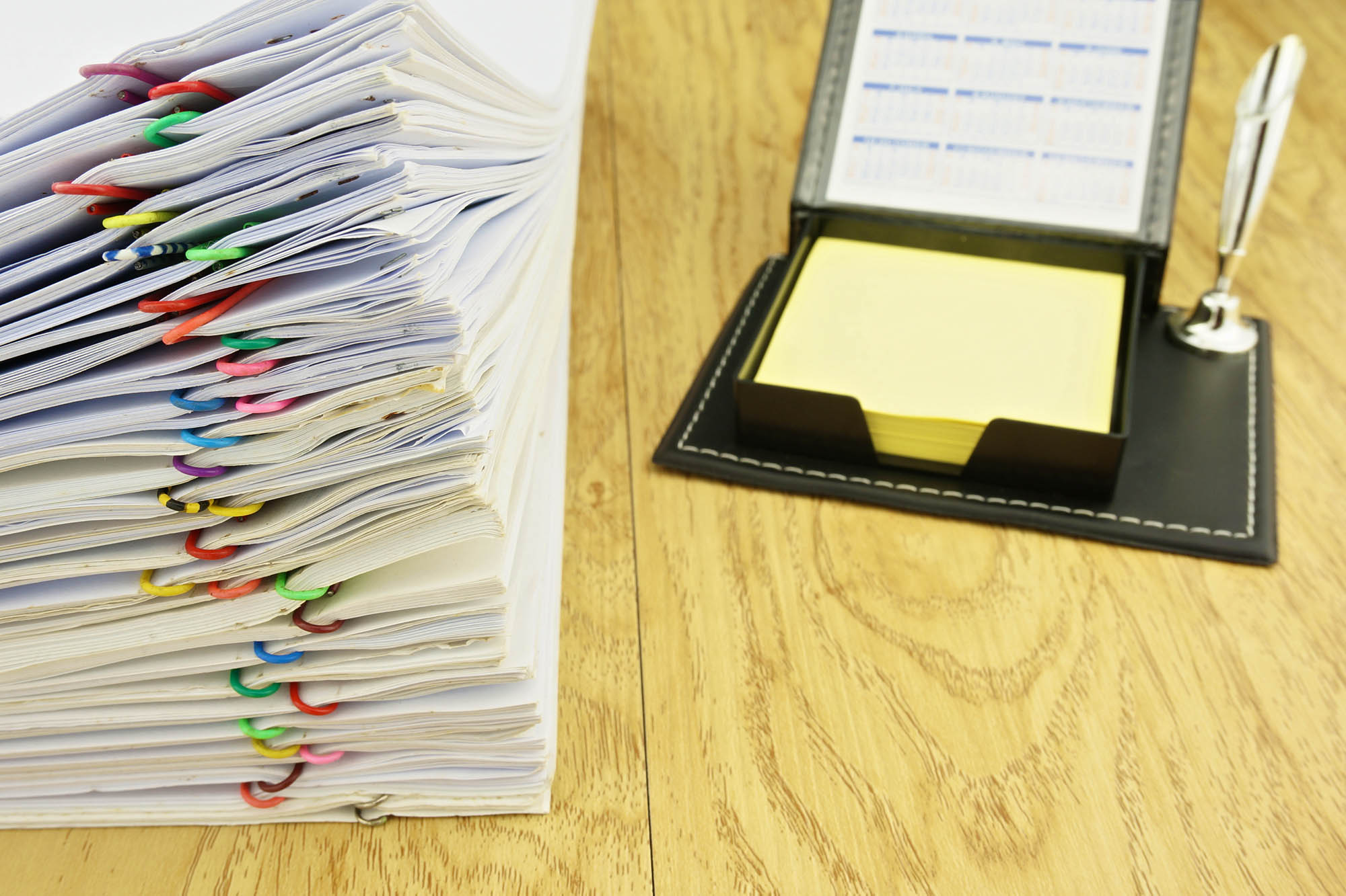 На столе лежат стопки документов с разноцветными кольцами, рядом стоит органайзер с календарем и желтыми листами, а также ручка в подставке.