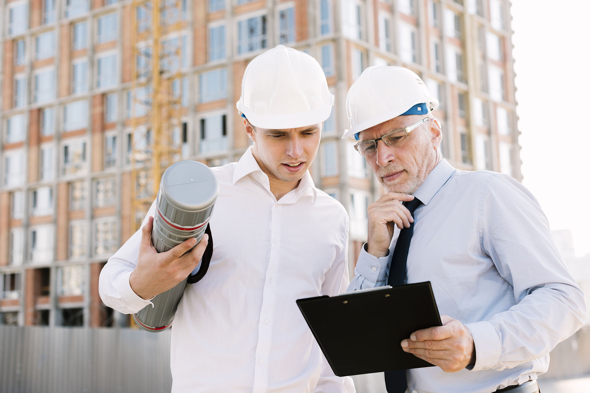 Два человека в строительных касках и очках рассматривают чертежи.